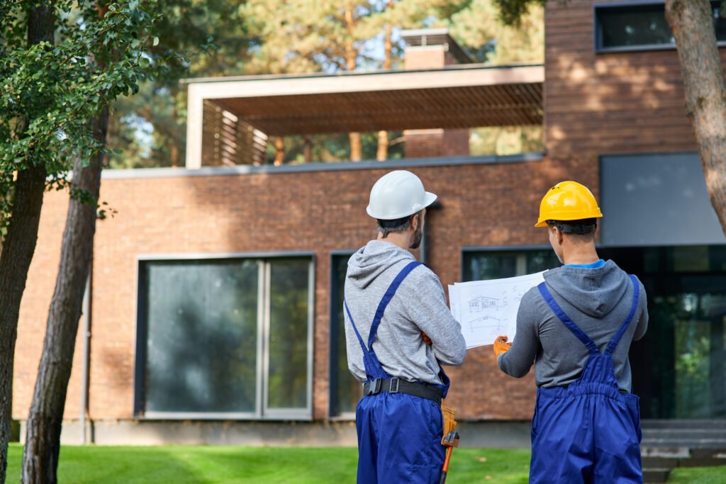 Rear view of two builders standing outdoors with an open blueprint discussing the half completed cottage house. Building, teamwork, partnership concept