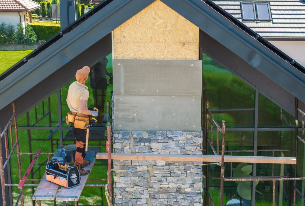 A construction worker is positioned on scaffolding, applying a stone facade to the upper section of a residential building. The work is taking place in a suburban area, with green grass and trees visible in the background, indicating a fresh, sunny day.
